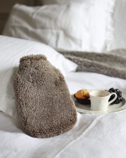 A brown sheepskin hot water bottle, shown on a bed with a plate of food in the background