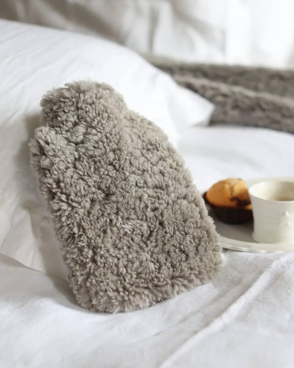 Pale grey sheepskin hot water bottle on a white bed with a blurred background.