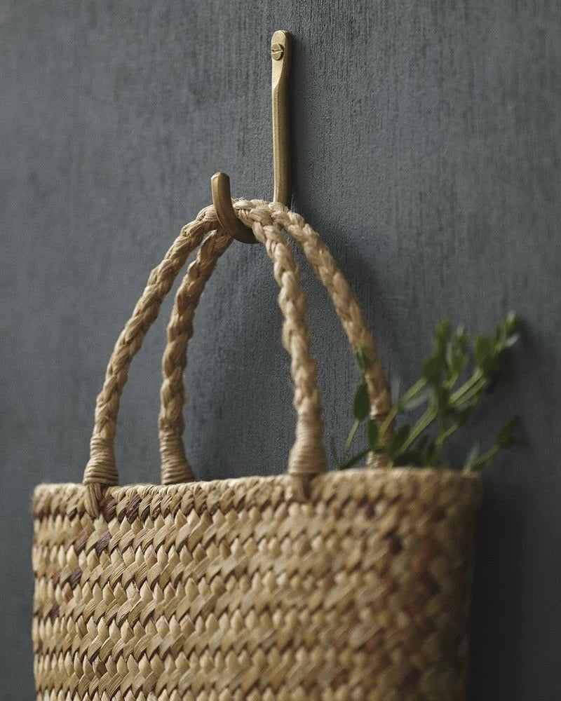 A brass wall hook, with a woven basket, hanging on a hook against a dark wall.