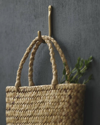 A brass wall hook, with a woven basket, hanging on a hook against a dark wall.