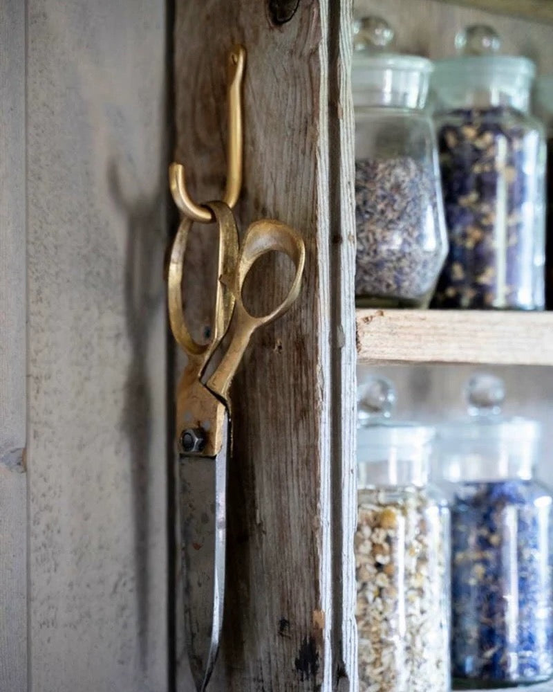 Brass scissors hanging on a brass hook, wall with jars of herbs in the background