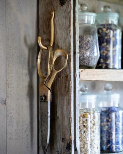 Brass scissors hanging on a brass hook, wall with jars of herbs in the background