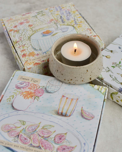 Lit tealight candle in ceramic holder on top of illustrated tealight boxes