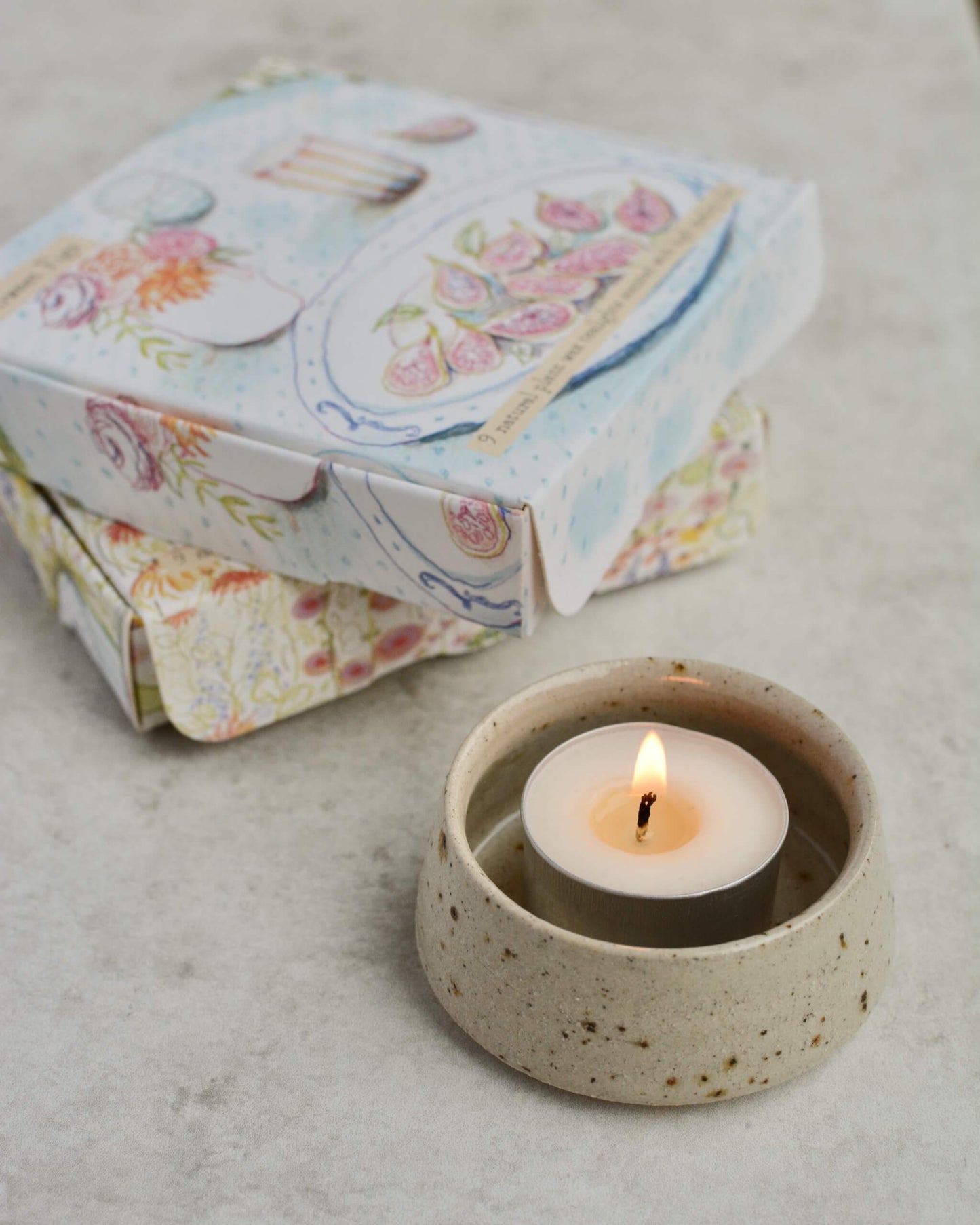 Lit tealight candle in ceramic holder in front of illustrated tealight boxes