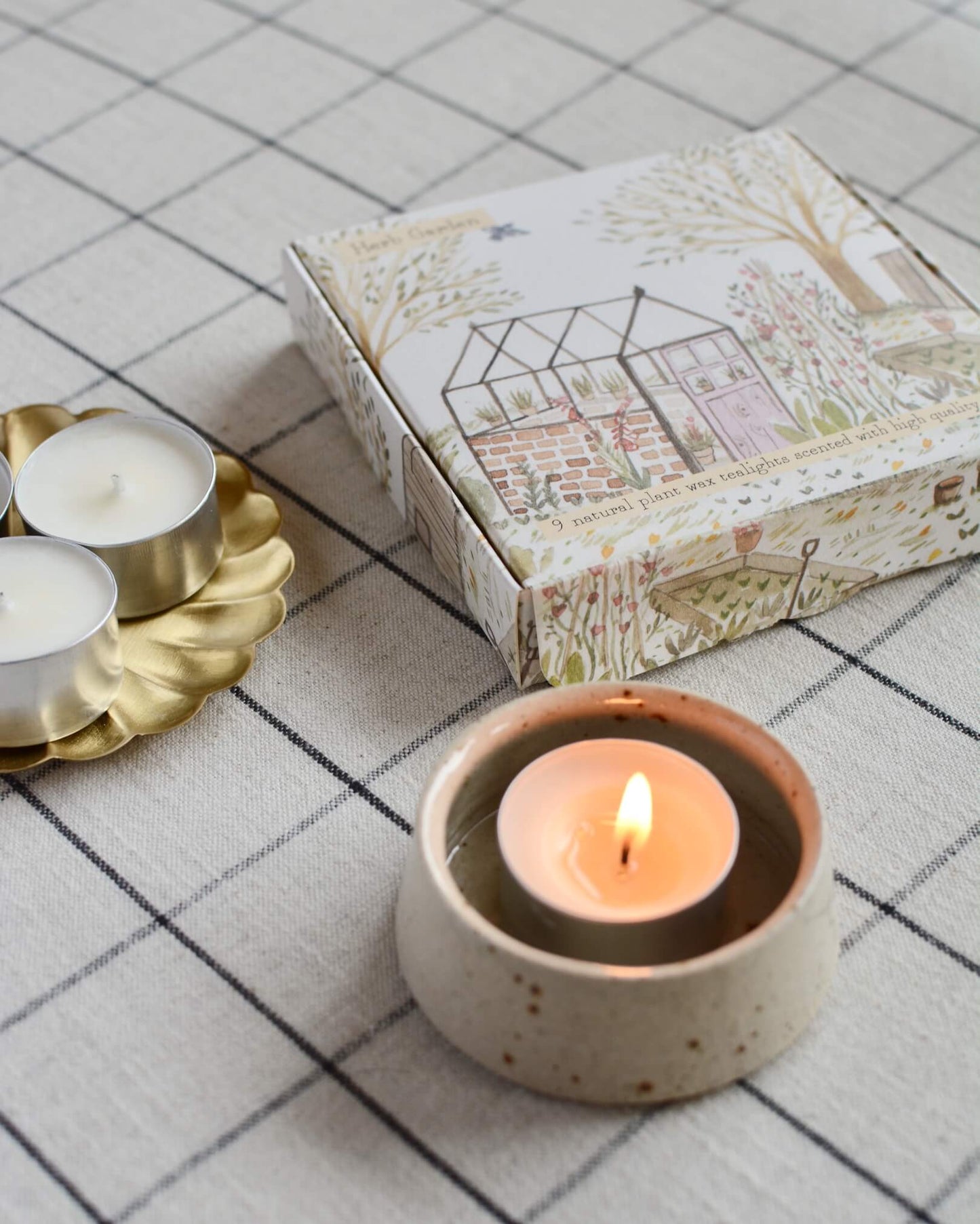 A lit tea light next to a box of tealights, with an illustrated box featuring a glasshouse within a summer garden.