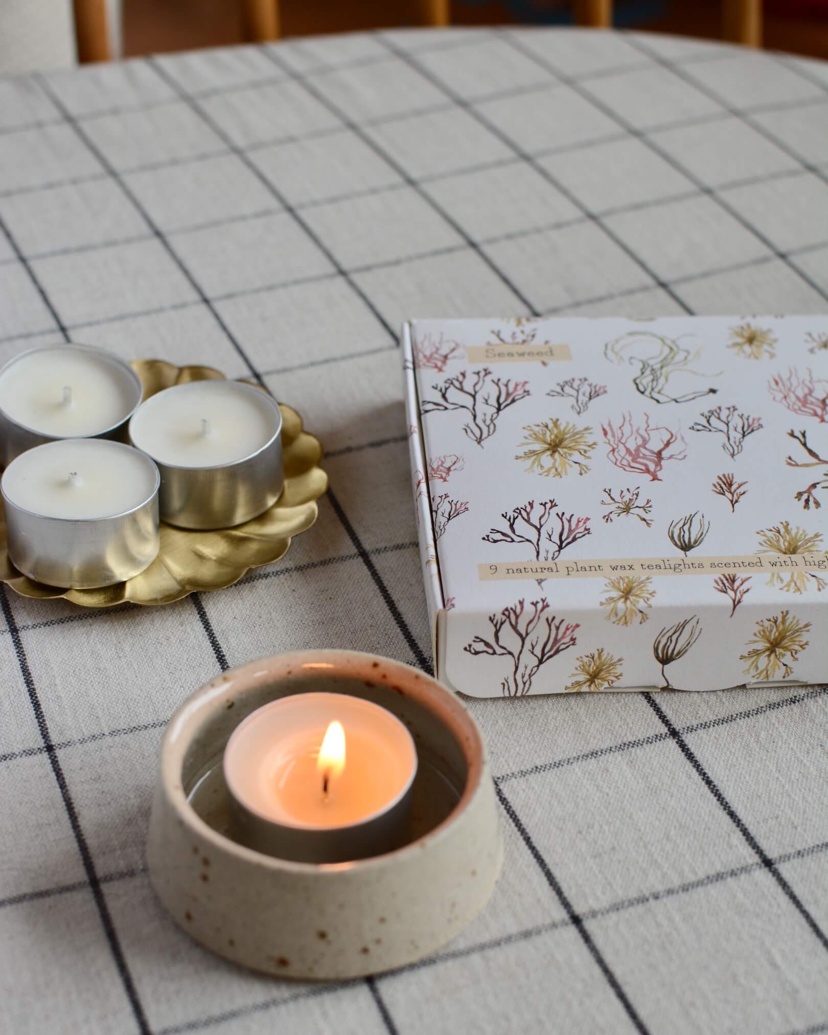 A lit tea light in a ceramic holder, next to an illustrated box of tea lights featuring watercolour illustrations.