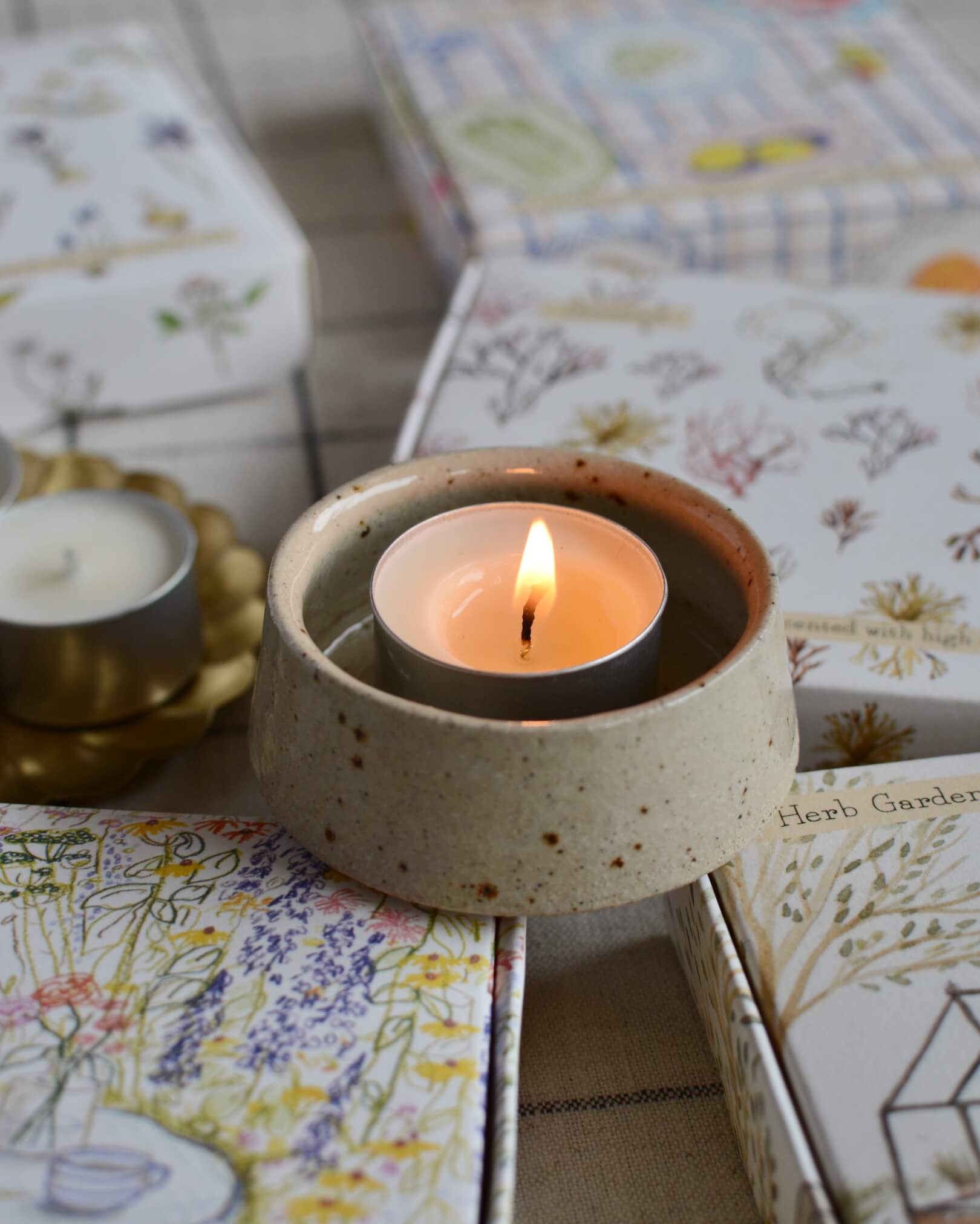 A lit tea light in a ceramic holder, on top of a collection of illustrated tea light boxes.