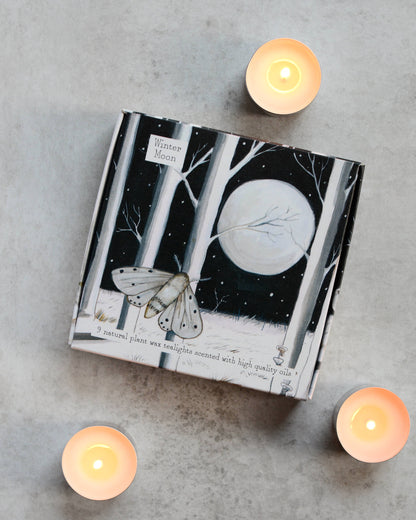 A set of lit tealights alongside a box with a monochrome forest and moon illustration.