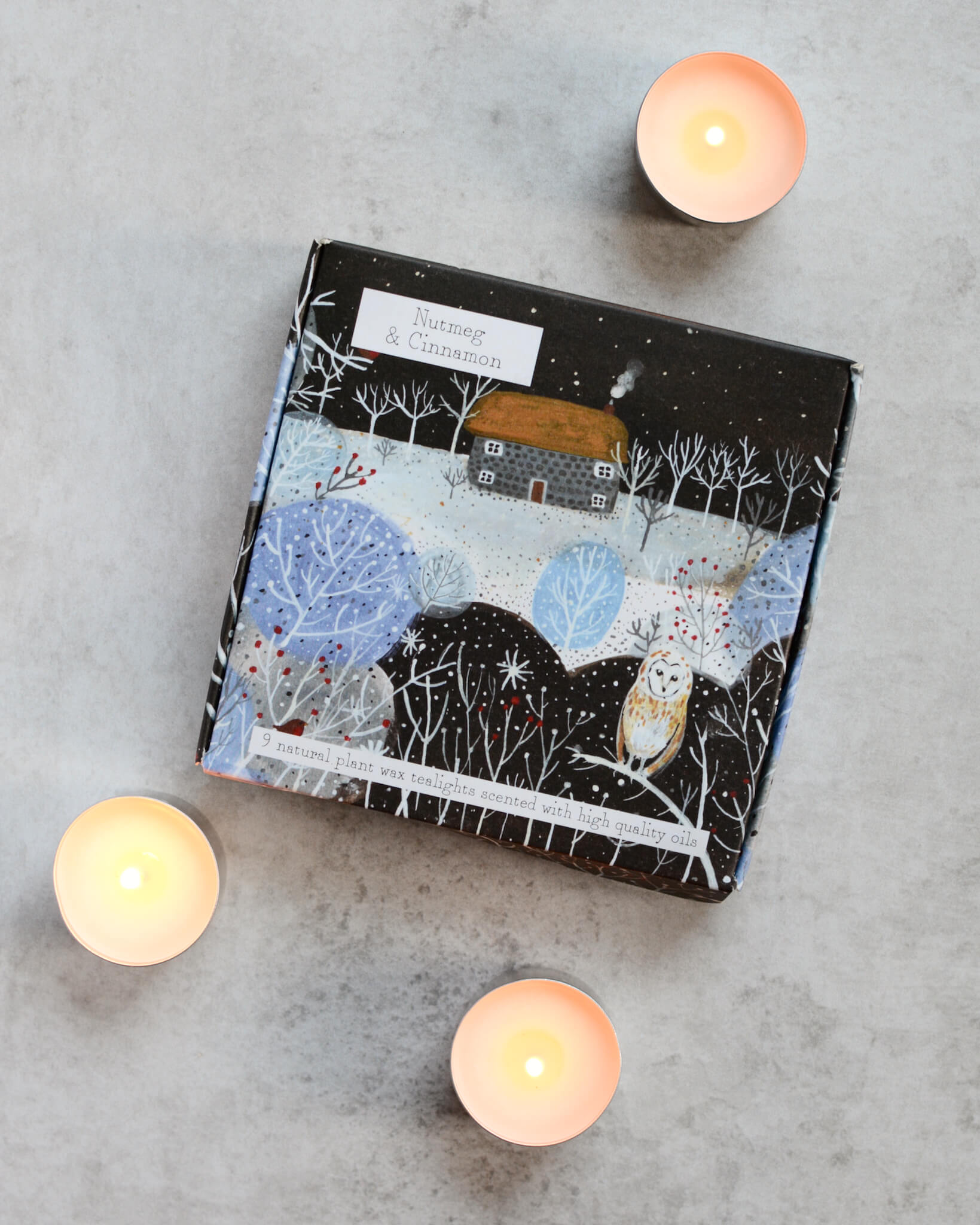A set of tealights with a Nutmeg and Cinnamon scent, displayed alongside their packaging which features a wintery landscape illustration.