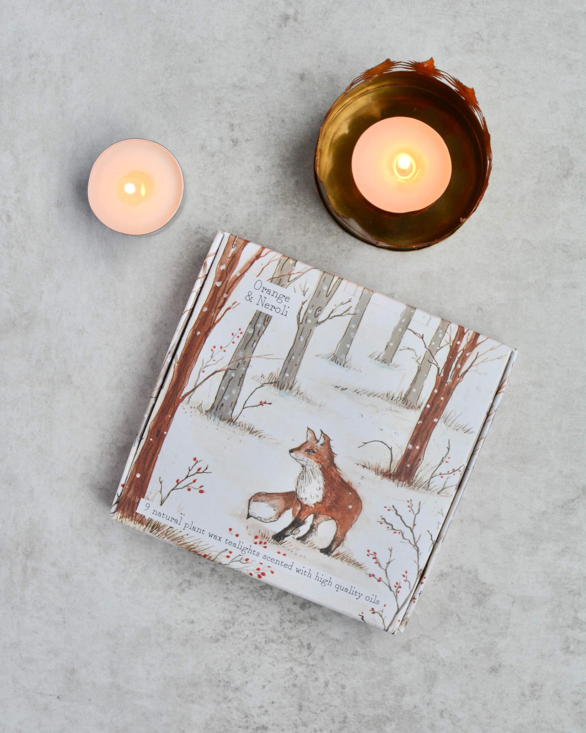 A box of tealights with fox illustration between two lit candles on a grey surface