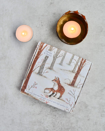 A box of tealights with fox illustration between two lit candles on a grey surface