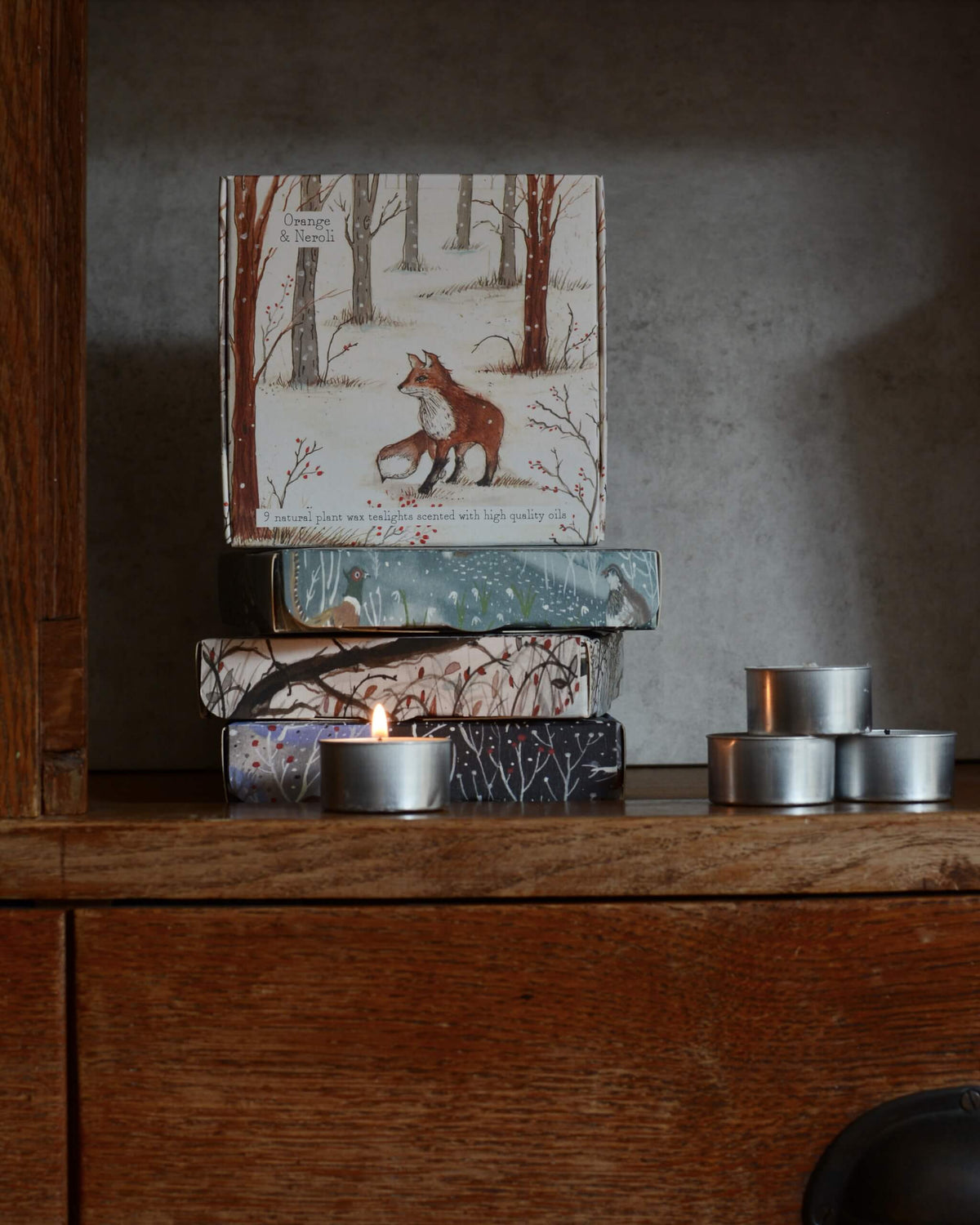 A stack of decorative tealight boxes with winter forest illustrations sits on a wooden shelf beside several silver tea light tins, including one lit candle, against a textured gray wall.