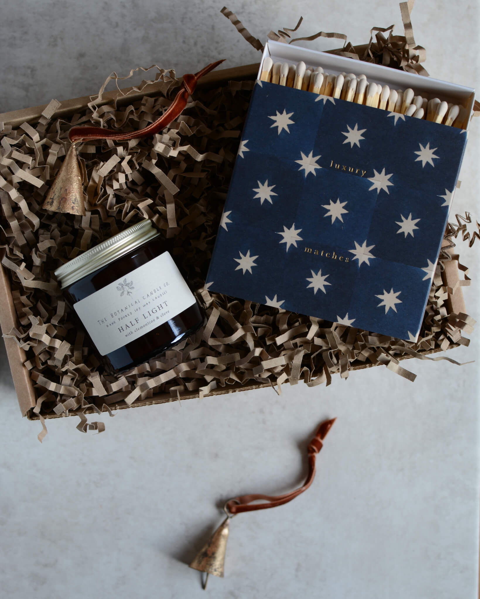 A Christmas gift box containing a glass jar festive candle, a box of matches with a star design and a pair of brass bell ornaments.