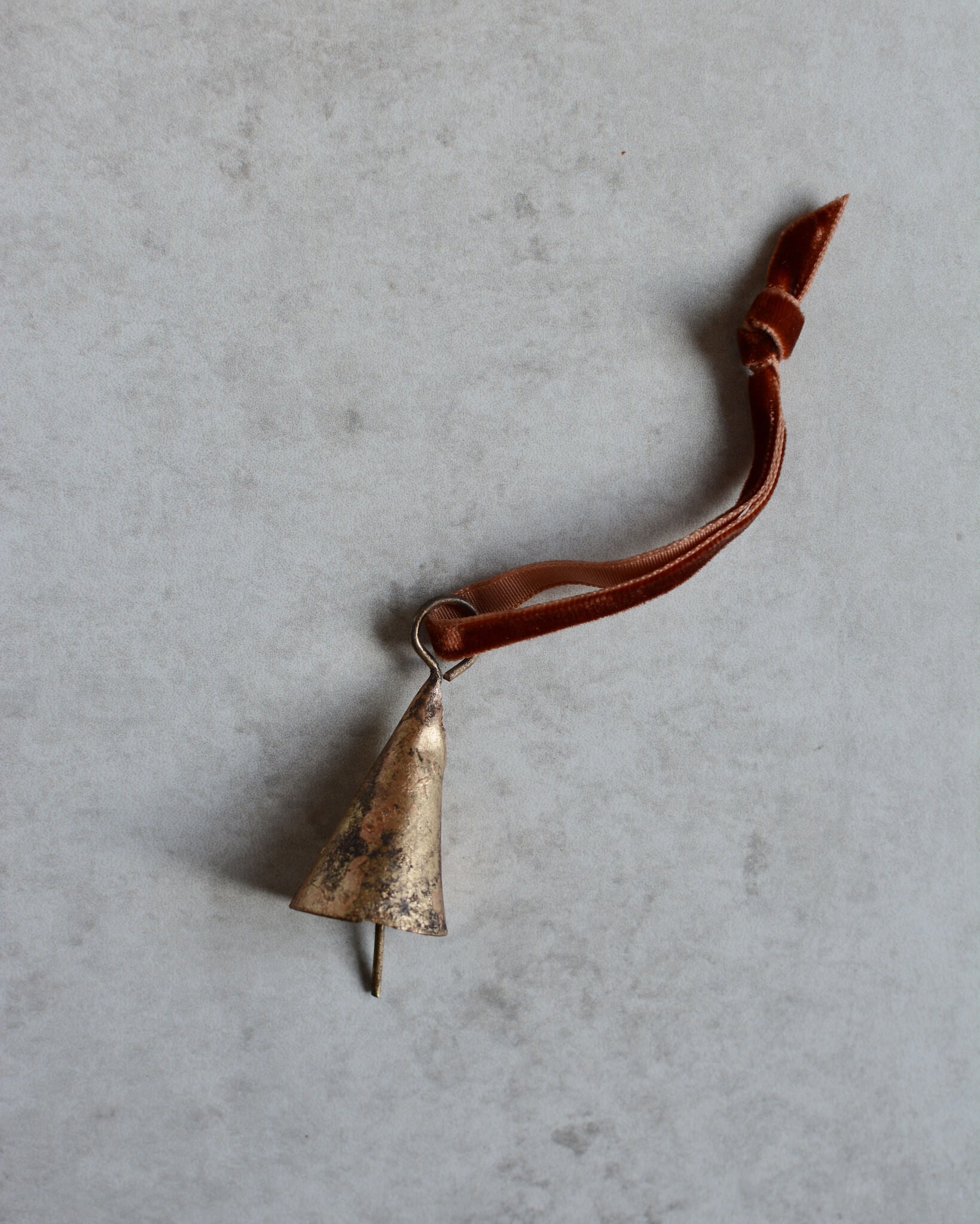 A brass bell ornament, strung onto a rust orange velvet ribbon.