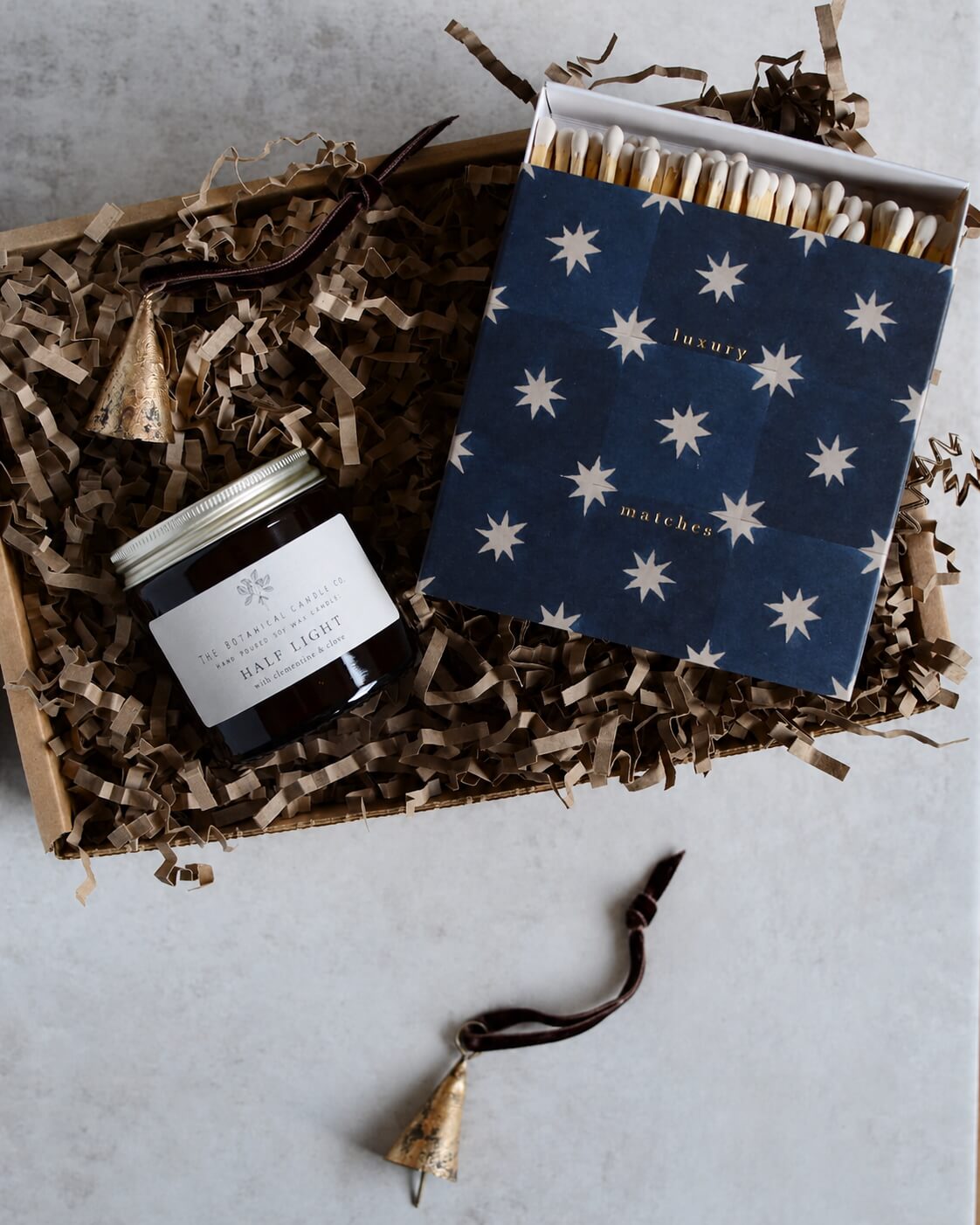 A candle gift set, featuring a navy blue matchbox with white star pattern labelled 'luxury matches' and a glass jar candle and a pair of brass bell ornaments.