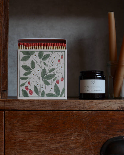 A square box of matches featuring an illustration of wild strawberries, alongside a candle in a glass jar.