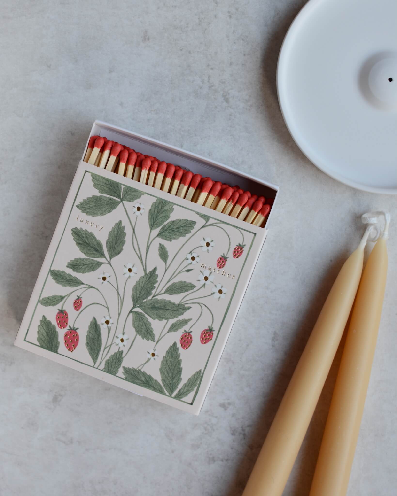 A square box of matches featuring an illustration of wild strawberries, alongside a pair of beeswax candles and an incense holder.