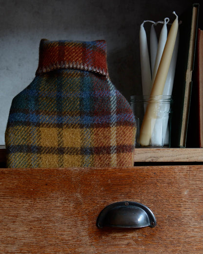 Plaid wool hot water bottle on a wooden surface with candles in the background
