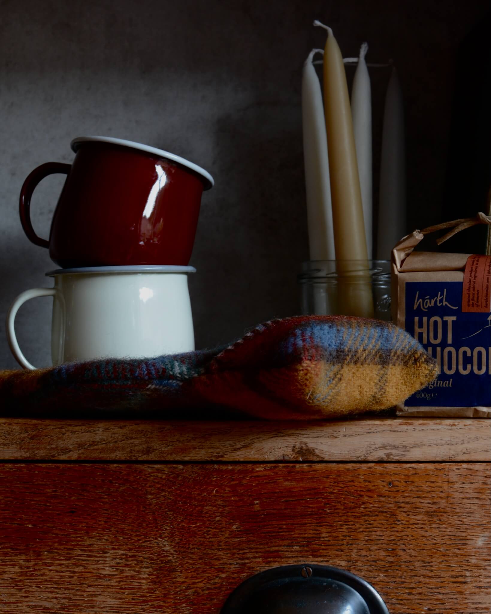 Two enamel mugs stacked on a wooden surface with candles, hot chocolate and a hot water bottle, in the background.