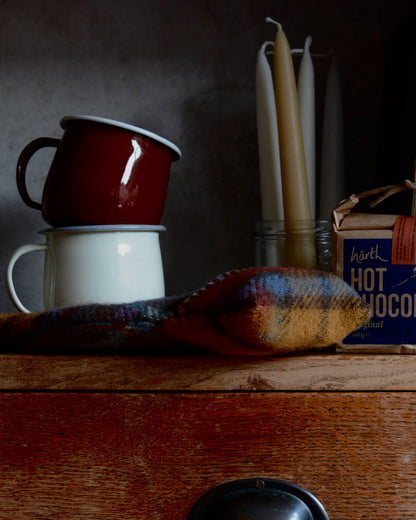 Two enamel mugs stacked on a wooden surface with candles, hot chocolate and a hot water bottle, in the background.