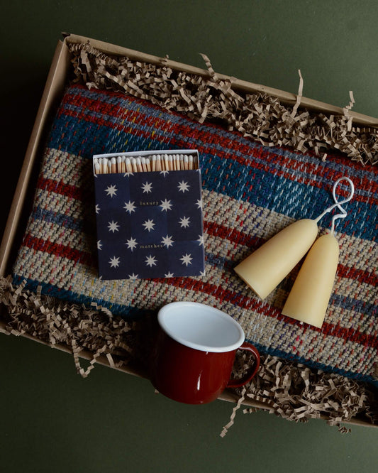 A gift box containing a wool blanket, box of matches, red enamel mug and pair of stubby beeswax candles.