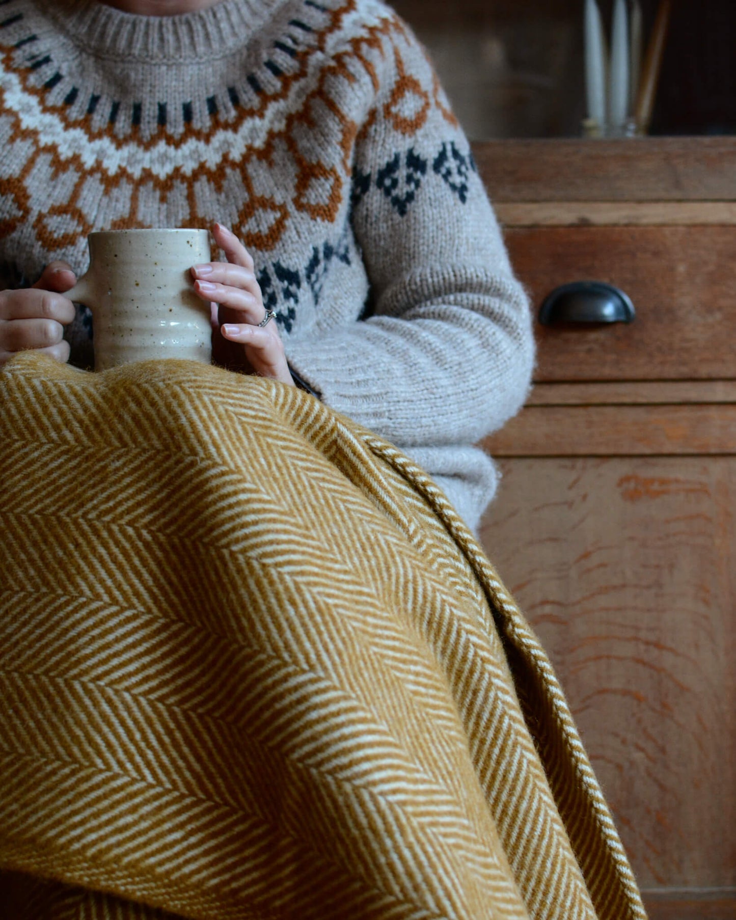 A woman with a mustard yellow wool herringbone blanket on her lap and a coffee cup.
