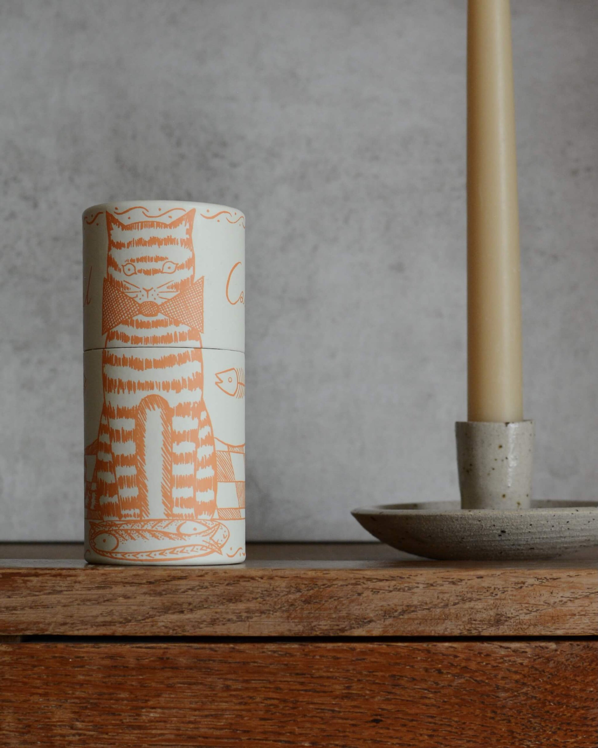 A cylindrical match box featuring an illustration of a cat with fish, on a wooden surface, with a candle holder to the side.