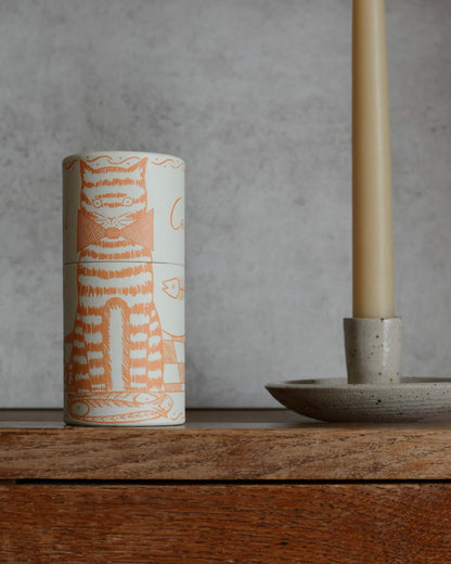A cylindrical match box featuring an illustration of a cat with fish, on a wooden surface, with a candle holder to the side.