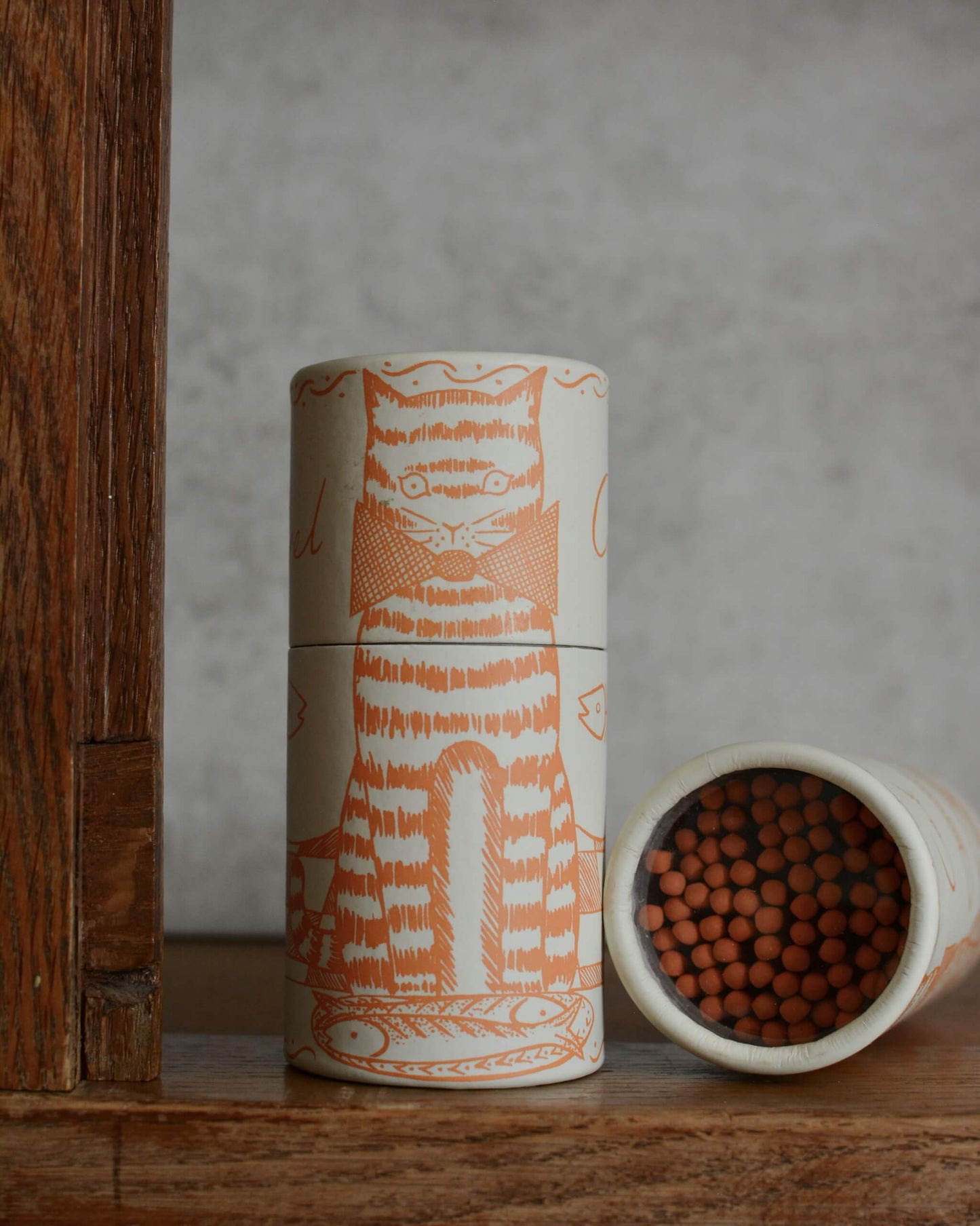 A cylindrical matchbox with a orange cat design, standing upright next to another matchbox tipped on its side showing the orange match tips.