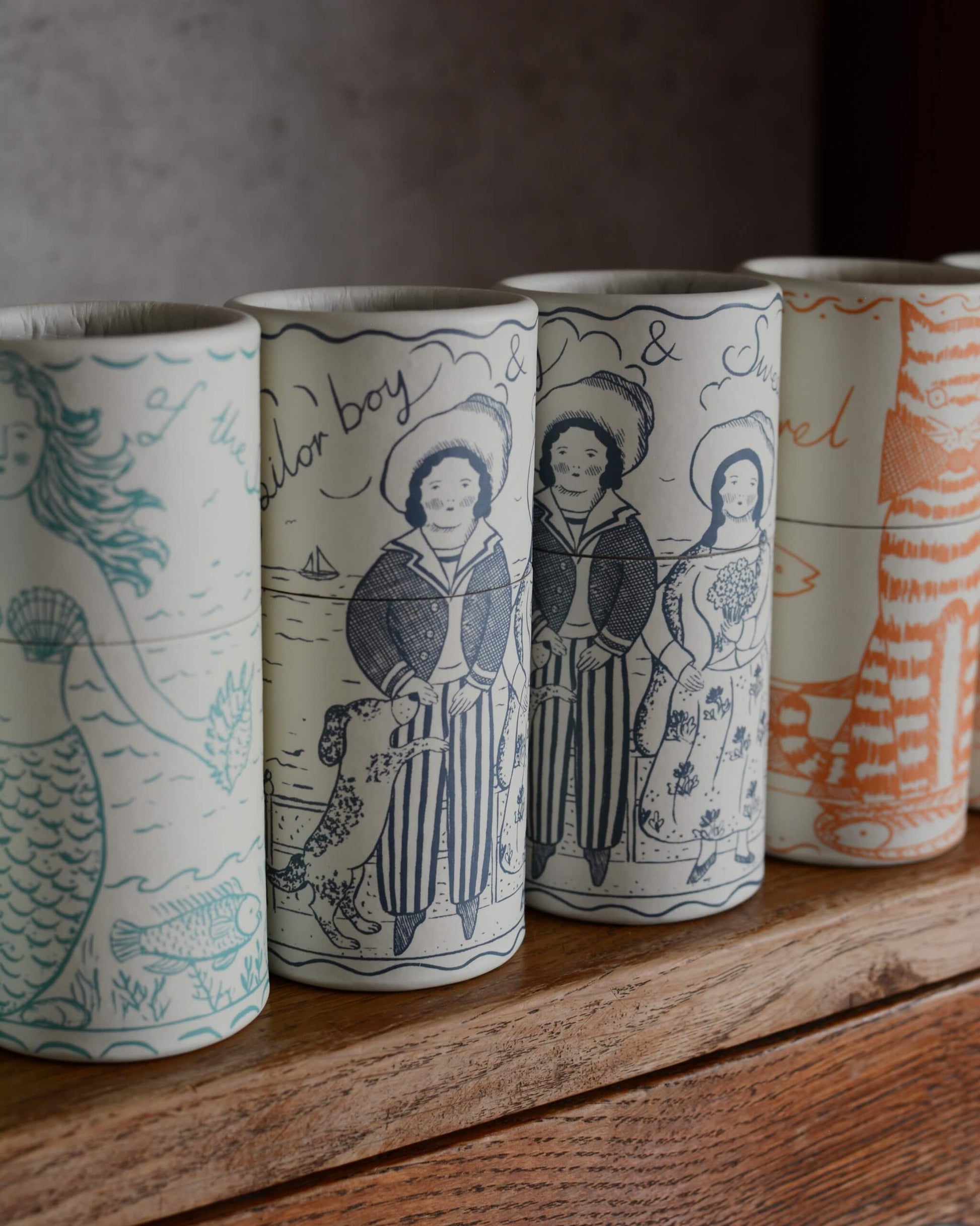 A row of cylindrical illustrated match tubes with whimsical illustrations, including a mermaid, a sailor boy, and a cat.