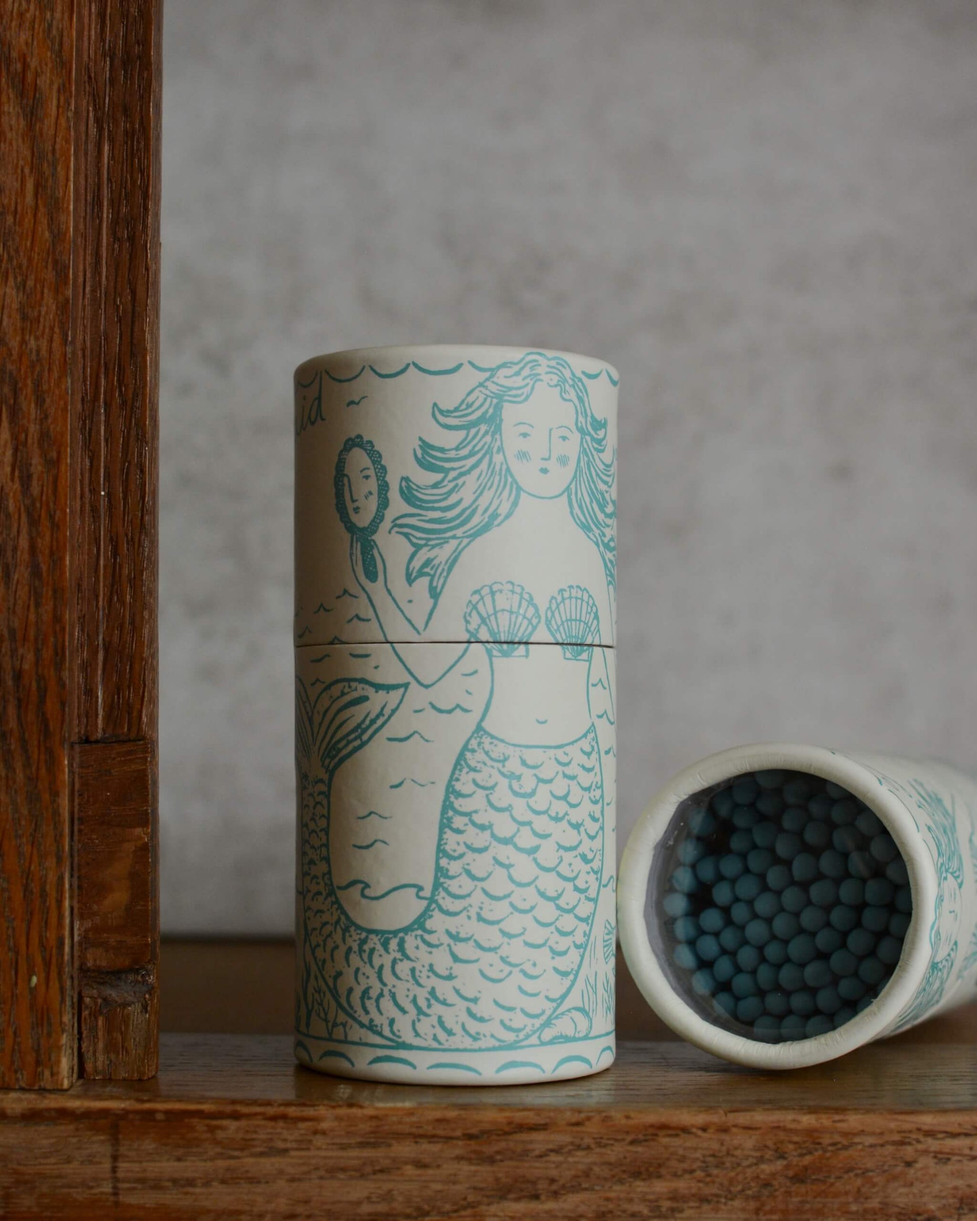 Image of a cylindrical matchbox featuring a mermaid illustration, next to the same matchbox on its side showing the blue match tips.
