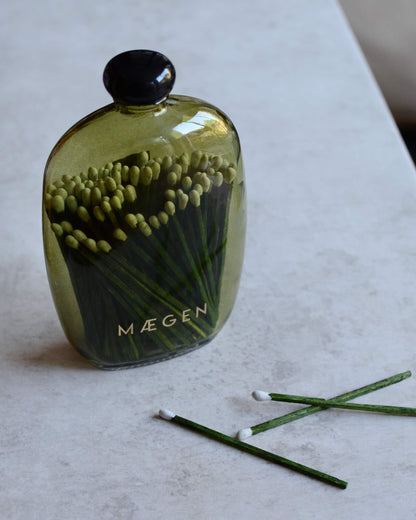 Olive green glass matches bottle with a black cap, with the brand name 'MÆGEN' printed on the front. Three green matchsticks with white tips lie on a light stone surface in the foreground.