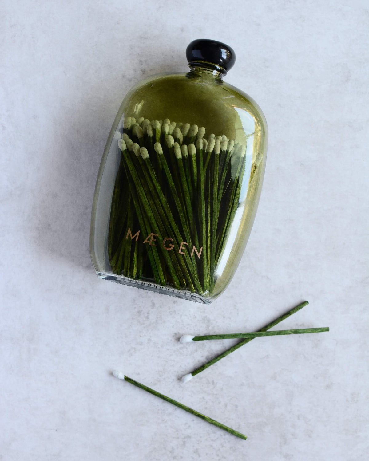 A green glass bottle with a black cap containing green matchsticks, with the brand name 'MÆGEN' printed on the front.