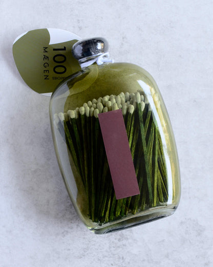 A clear glass bottle filled with green matchsticks, showing a striker on the reverse, with a green paper tag that reads '100 matches'.