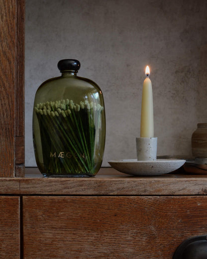 A dark olive green glass matches bottle with a black cap and gold lettering, sits on a rustic wooden shelf next to a lit beeswax taper candle in a speckled ceramic holder.