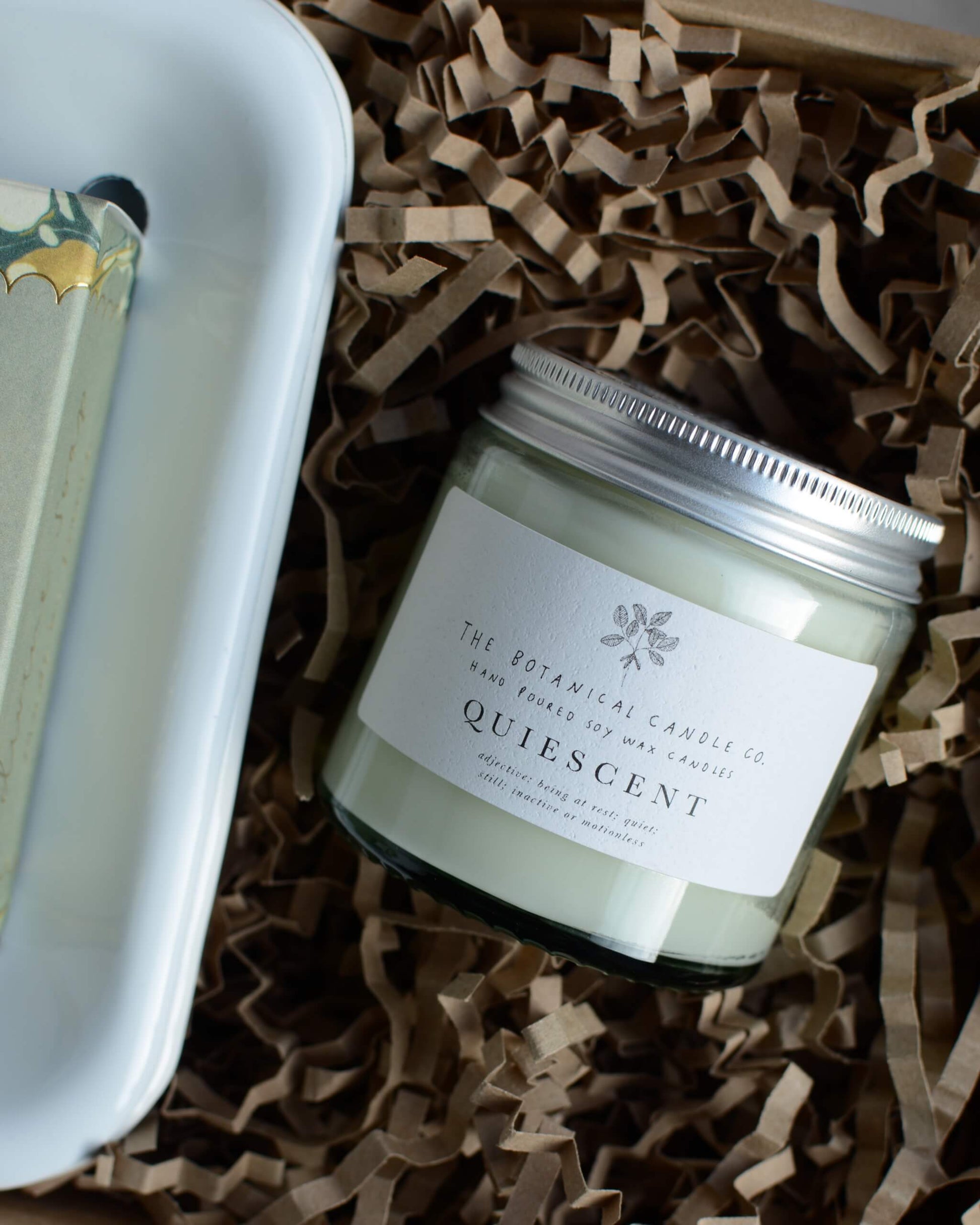 Candle jar labeled 'Quiescent' on crinkled brown paper in a gift box.