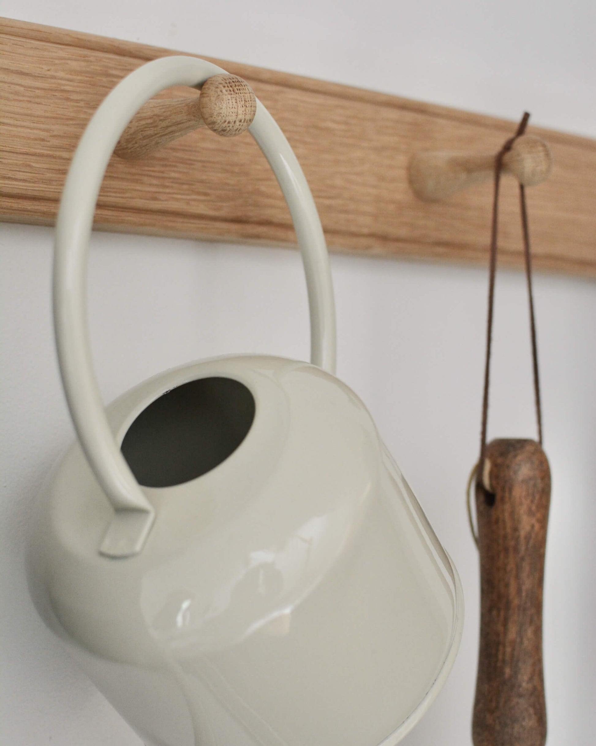 Cream enamel watering can and garden tool, hanging on an oak peg rail.
