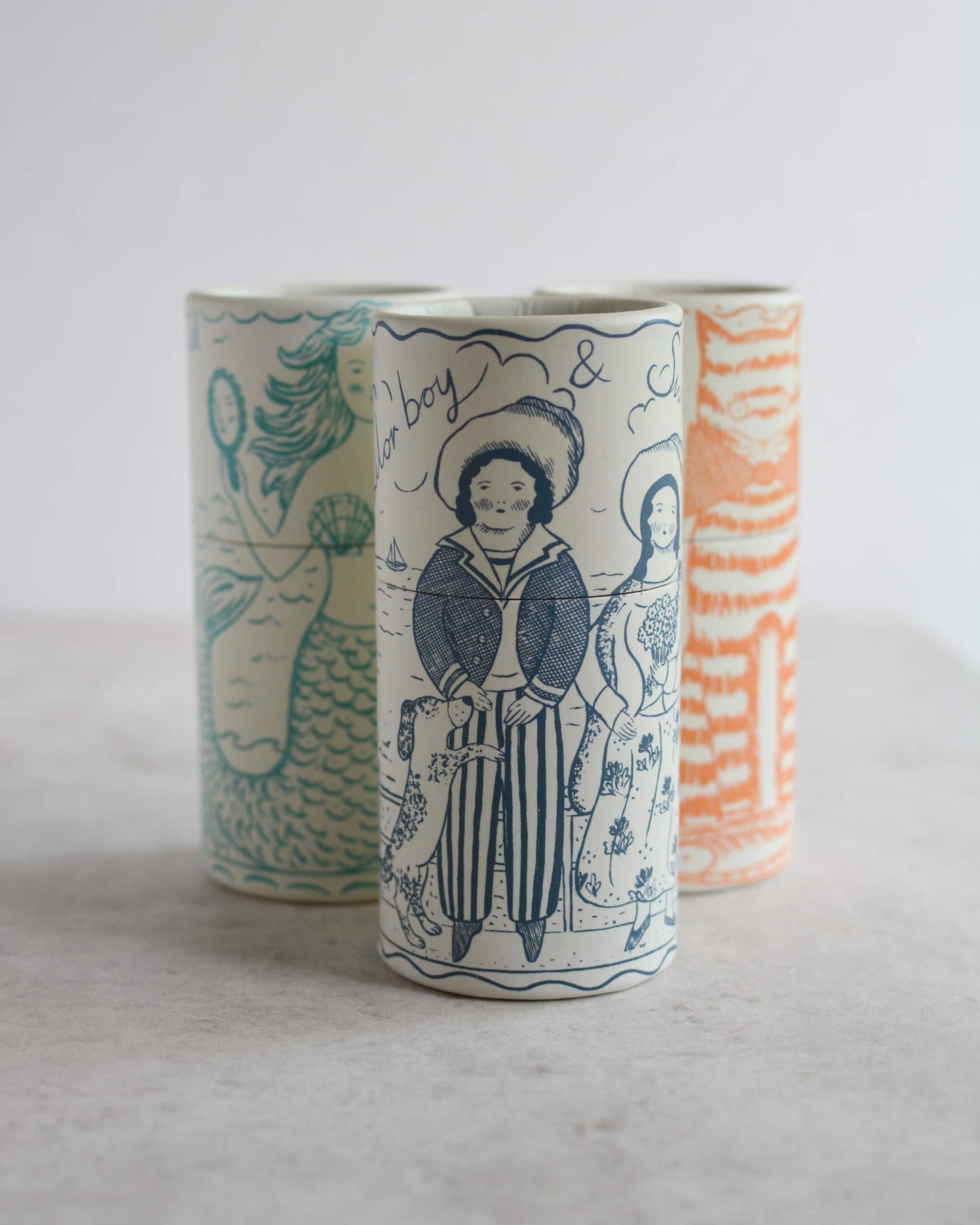 Three cylindrical illustrated match tubes with whimsical illustrations, including a mermaid, a sailor boy, and a cat, displayed on a light grey surface against a white background.