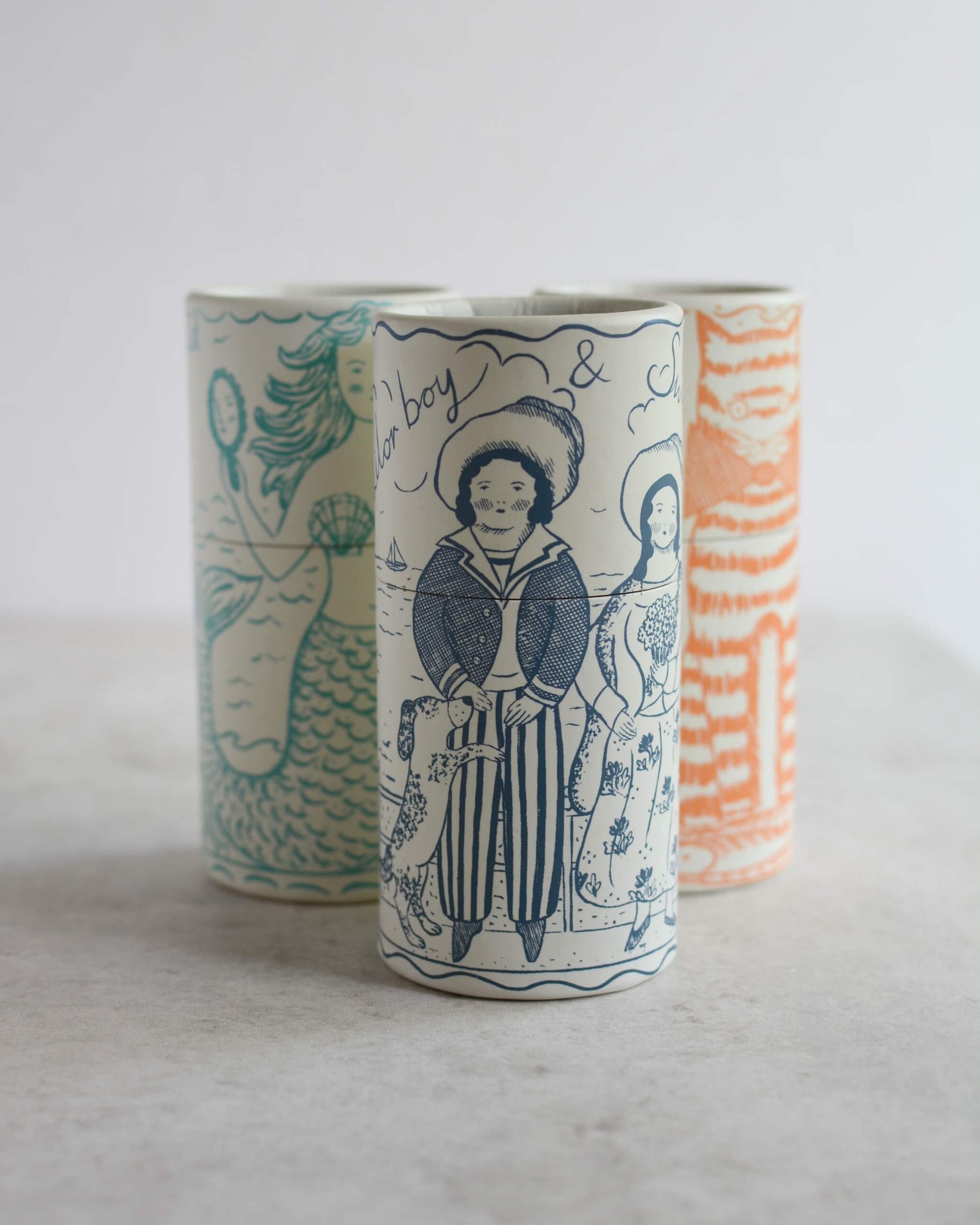 Three cylindrical illustrated match tubes with whimsical illustrations, including a mermaid, a sailor boy, and a cat, displayed on a light grey surface against a white background.