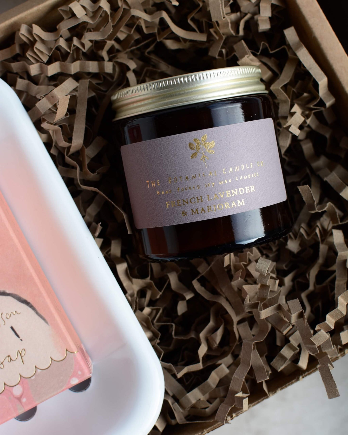 Candle jar with a label on brown crinkled paper, next to a pink soap bar.