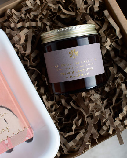 Candle jar with a label on brown crinkled paper, next to a pink soap bar.