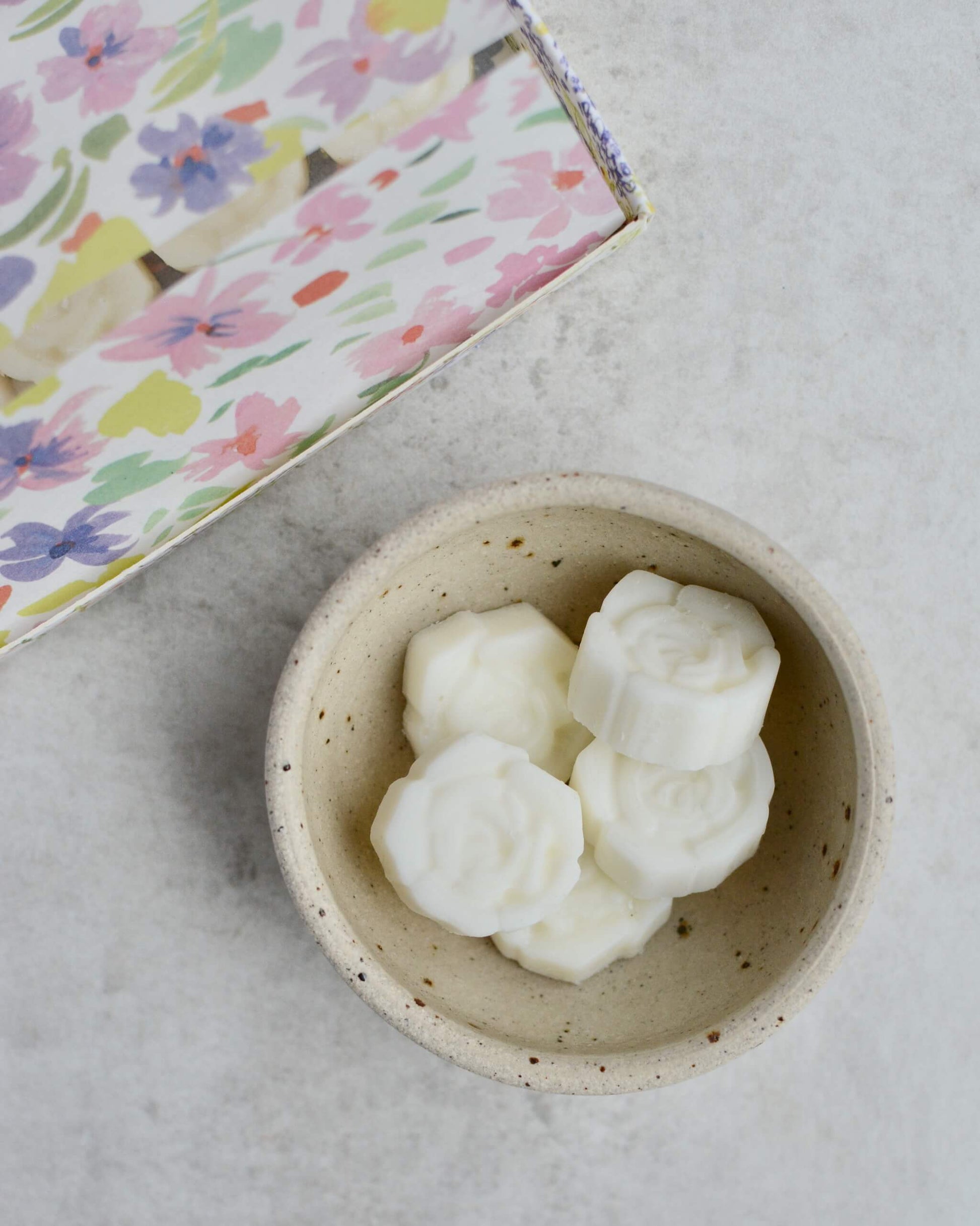 White flower-shaped wax melts in a small speckled bowl on a light grey surface with the floral-patterned box in the background.