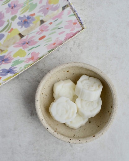White flower-shaped wax melts in a small speckled bowl on a light grey surface with the floral-patterned box in the background.