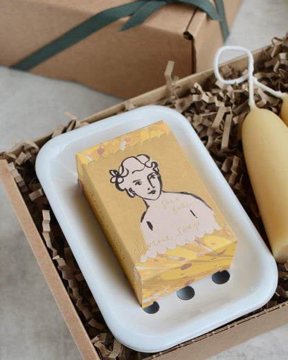Yellow soap bar with a decorative label on a white soap dish inside a gift box with brown paper.