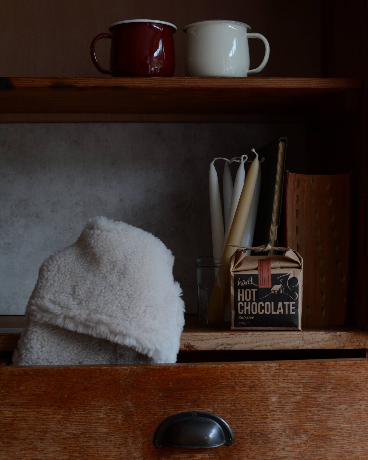 Two mugs on a shelf with a box of hot chocolate and a sheepskin hot water bottle in the foreground.