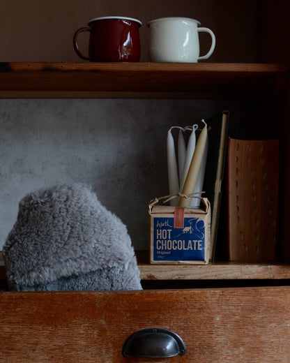 Two mugs on a shelf above a drawer with a box of hot chocolate and candles.