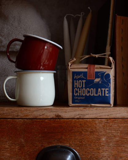 Two enamel mugs stacked on a wooden surface with a box of Harth Hot Chocolate nearby.