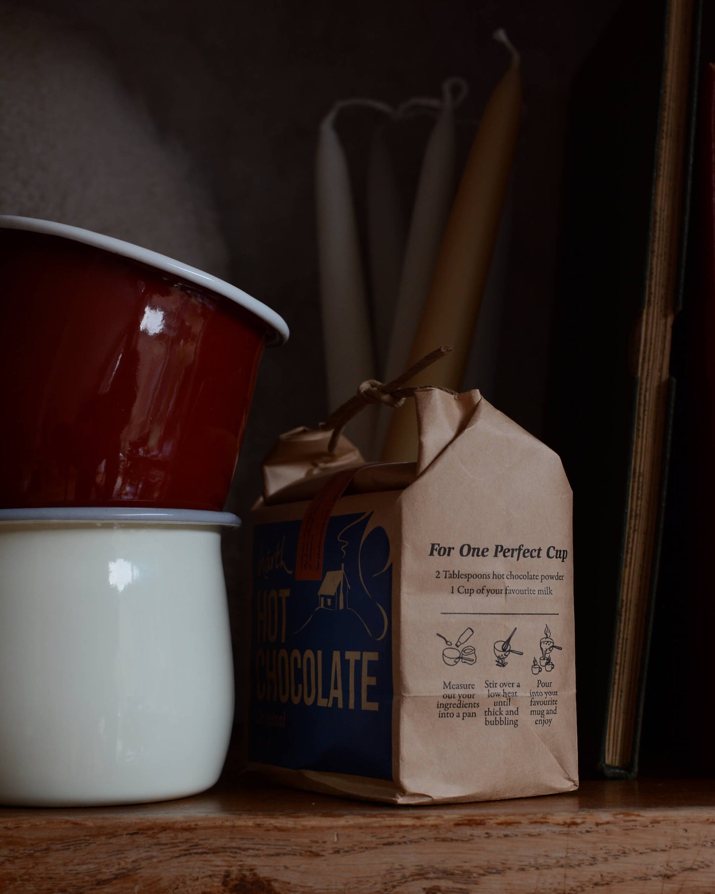 Two enamel mugs and a bag of hot chocolate on a wooden surface.