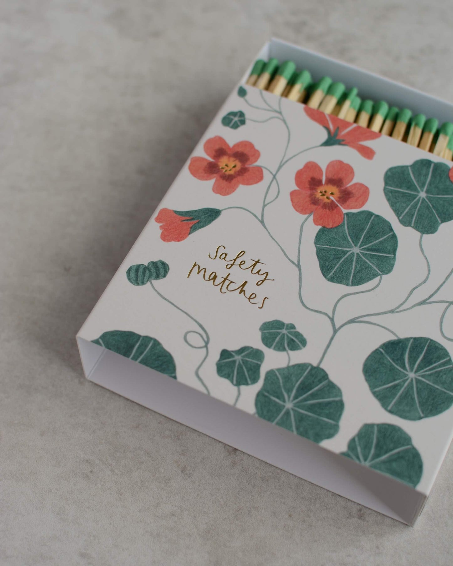 A decorative box of safety matches with a white cover printed with  pink nasturtium flowers and green leaves, shown open on a light grey stone surface.