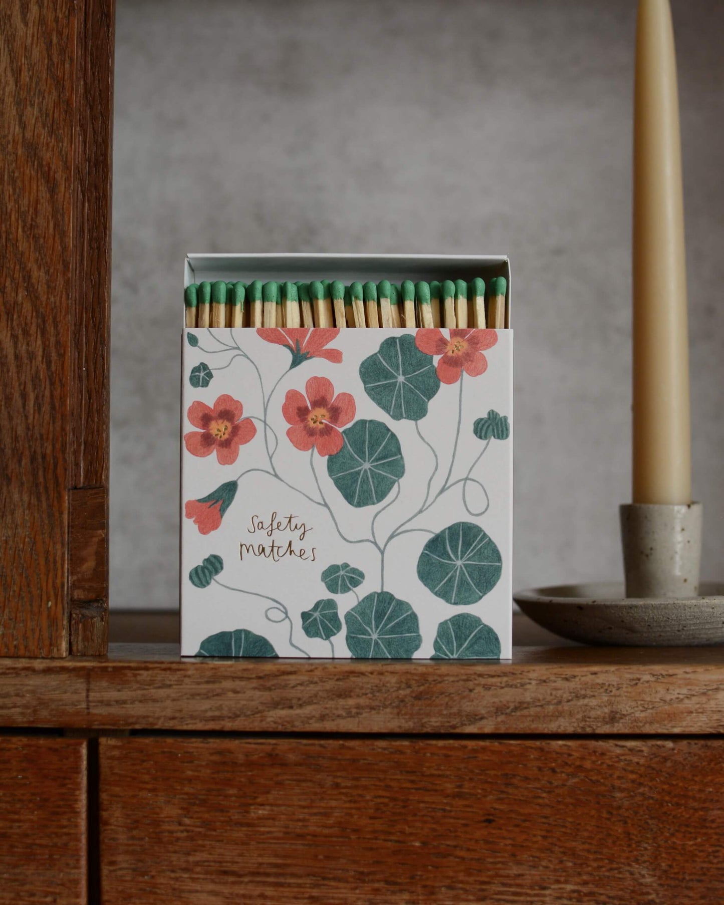 A decorative matchbox filled with safety matches sits on a wooden surface against a textured grey wall, with a candle holder visible on the right.